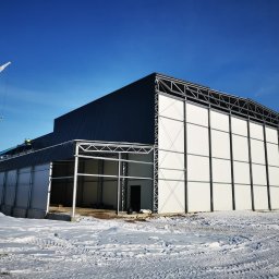 ELMET Elżbieta Jastrzębska - Montaż hali z płyt warstwowych w zimowej scenerii. Widoczny dźwig i pracownicy na wysokości. Częściowo ośnieżony teren budowy, błękitne niebo.