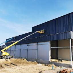 ELMET Elżbieta Jastrzębska - Montaż ciemnej elewacji hali z płyt warstwowych. Dwie platformy robocze z pracownikami na tle błękitnego nieba. Widoczne stalowe elementy konstrukcyjne i teren budowy.