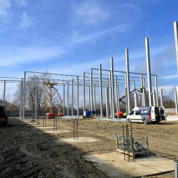 ELMET Elżbieta Jastrzębska - Budowa hali stalowej w trakcie montażu konstrukcji. Widoczne słupy, dźwigi oraz ekipa montażowa. Na pierwszym planie teren budowy, w tle błękitne niebo z chmurami.