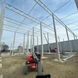 ELMET Elżbieta Jastrzębska - Wznoszenie szkieletu hali stalowej z użyciem podnośnika. Widoczne betonowe słupy i metalowe elementy konstrukcyjne. Częściowo ukończona budowa, teren budowy.