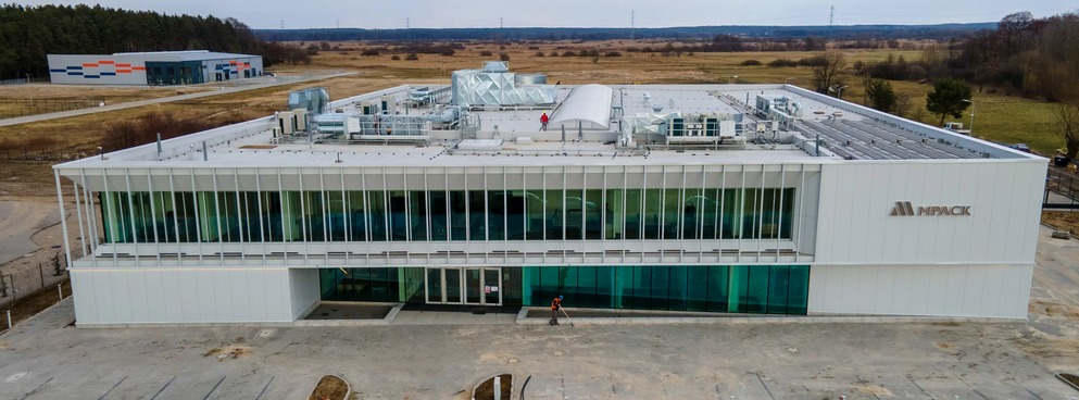 Nowoczesna hala z przeszkloną fasadą i białym wykończeniem, widok z góry. Na dachu instalacje i osoba w czerwonej kurtce. W tle krajobraz.