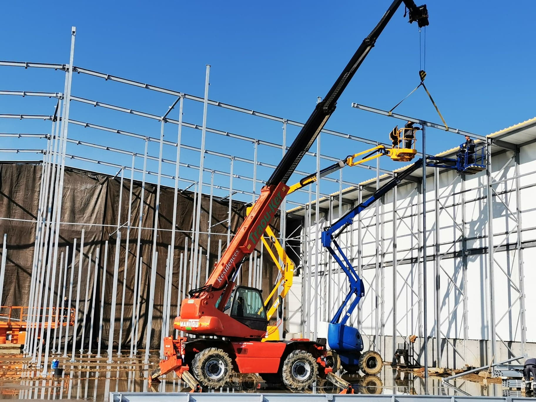 Montaż konstrukcji stalowej hali z użyciem podnośników. Widoczne stalowe elementy konstrukcyjne i pracownicy na podnośnikach. Częściowo zamontowana ściana hali w tle.