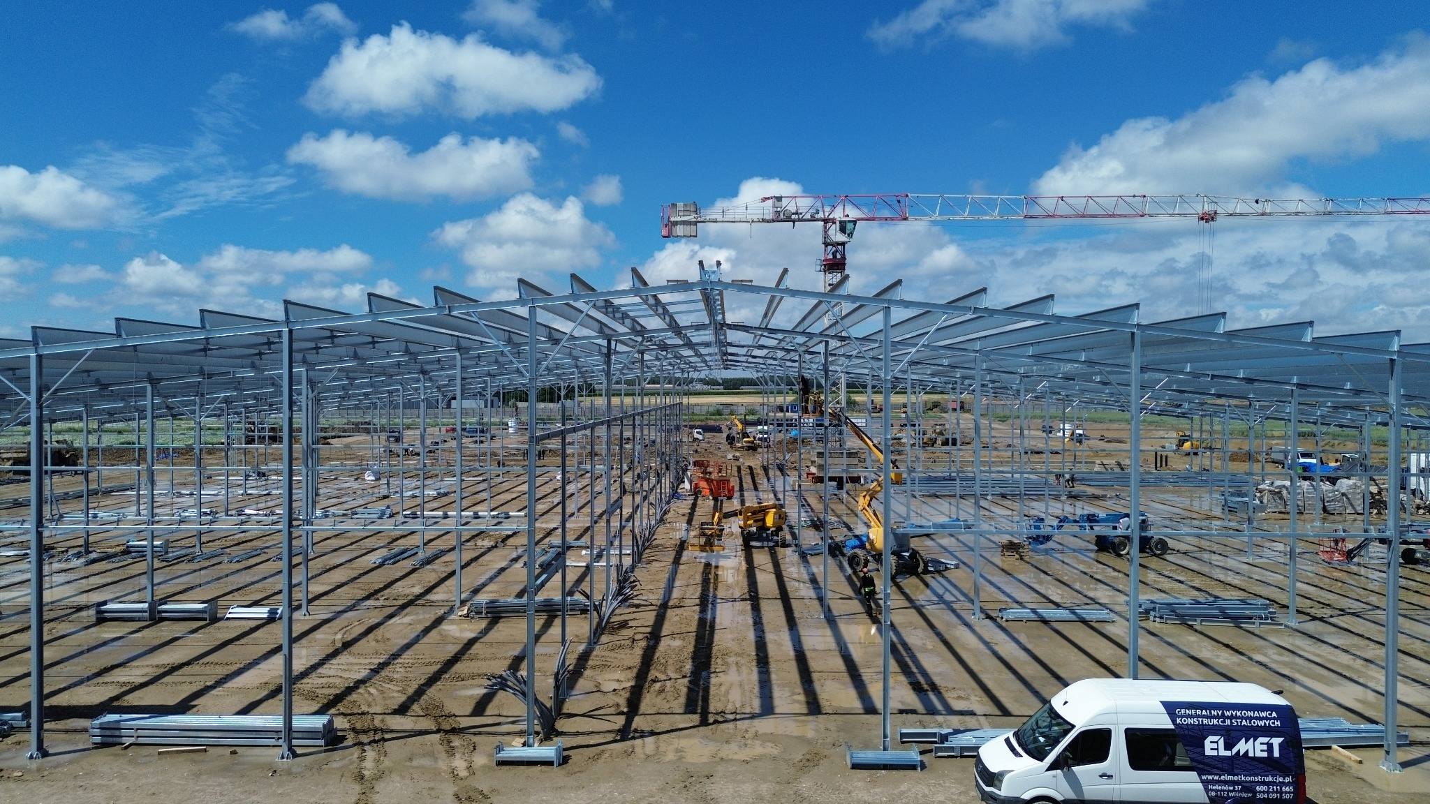 Konstrukcja stalowa hali w trakcie budowy, widok z góry. Na pierwszym planie biały bus firmy Elmet. W tle niebo z chmurami i żuraw budowlany.