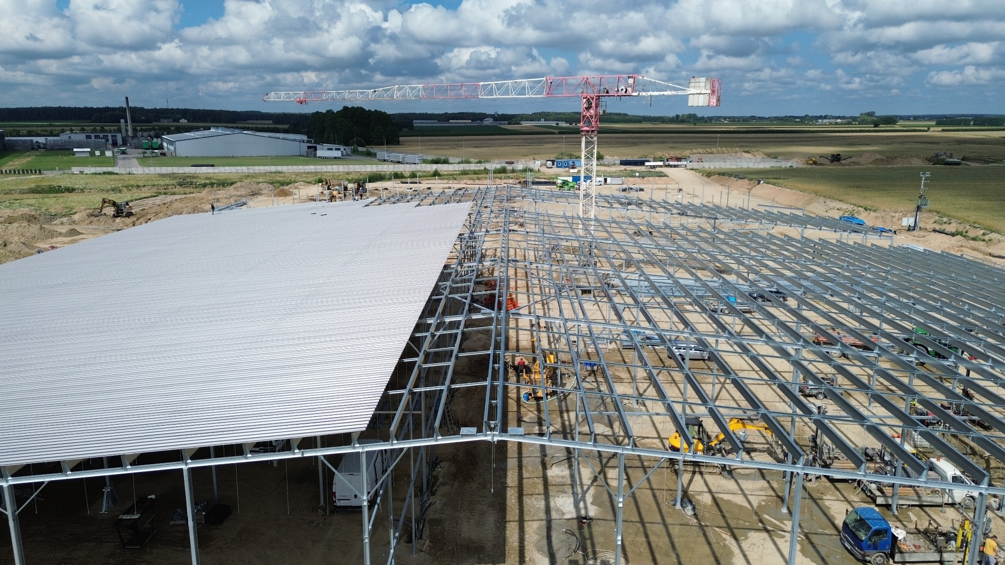 Budowa hali: widok z góry na stalową konstrukcję i częściowo zamontowane płyty warstwowe. W tle żuraw budowlany i krajobraz.