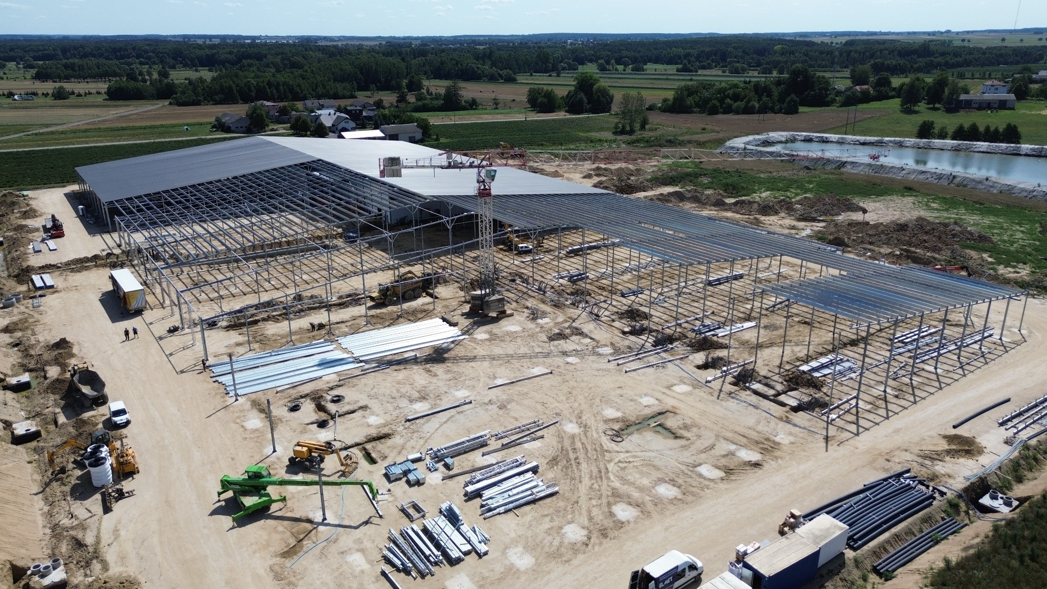Szeroki kadr z drona ukazuje budowę hali stalowej. Widoczne elementy konstrukcyjne, częściowo zamontowane płyty dachowe, sprzęt budowlany i materiały. W tle pola uprawne i zbiornik wodny.