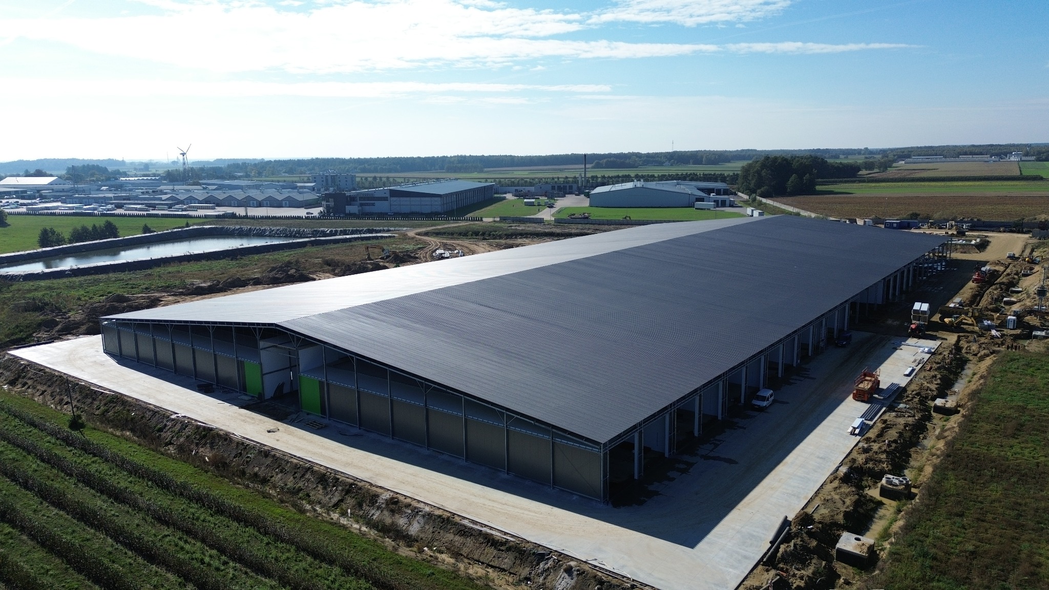 Szeroki kadr z lotu ptaka na halę z ciemną, płaską połacią dachu i metalową konstrukcją, obok widoczne prace ziemne i krajobraz z innymi budynkami w oddali.