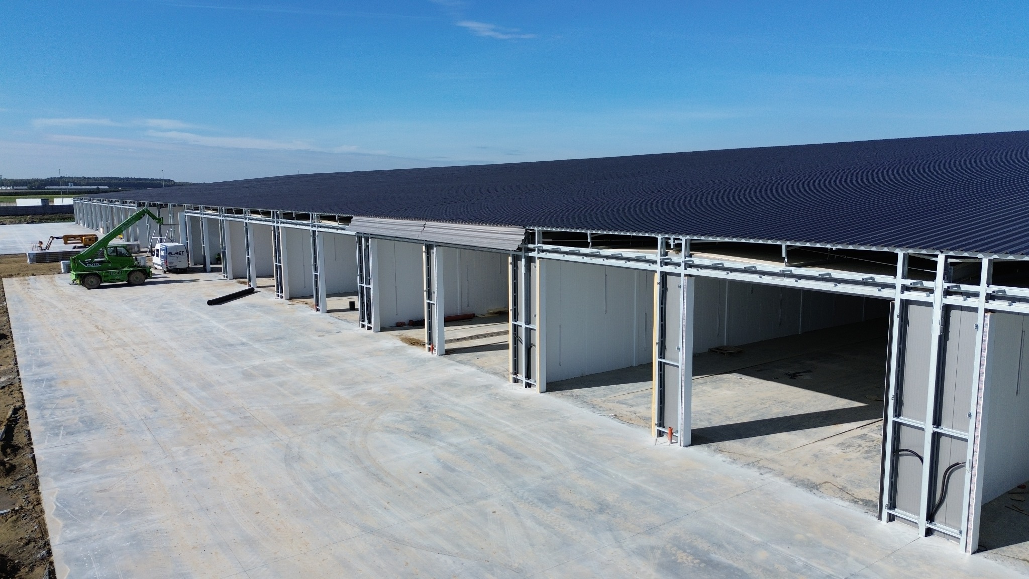 Hala w budowie z widocznym dźwigiem i elementami konstrukcyjnymi, oświetlona jasnym światłem słonecznym, na tle błękitnego nieba. Widok z góry.