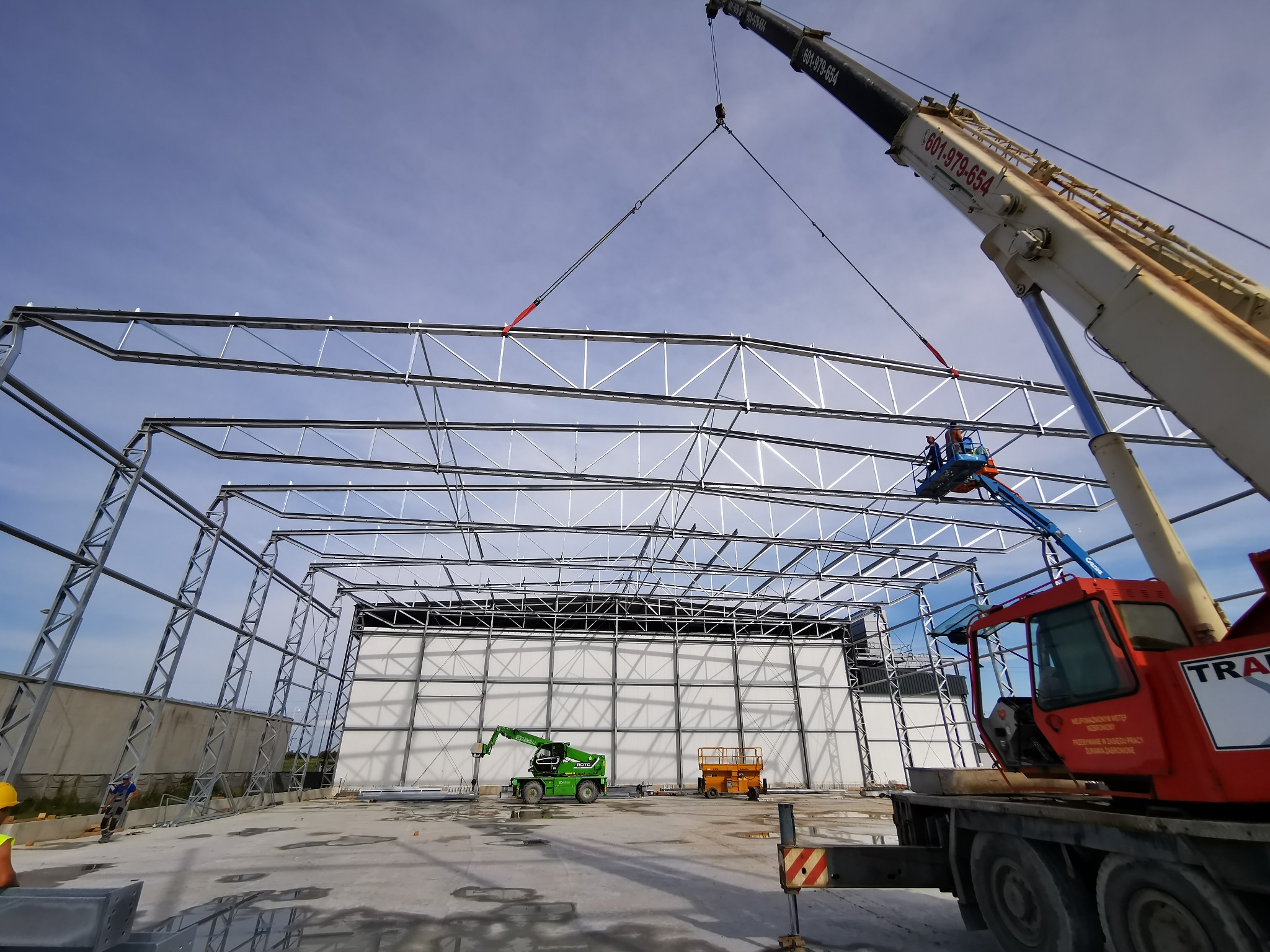 Montaż stalowej konstrukcji hali z użyciem dźwigu i podnośnika koszowego. Widoczne elementy konstrukcyjne, maszyny budowlane i pracownicy w tle. Jasne niebo.