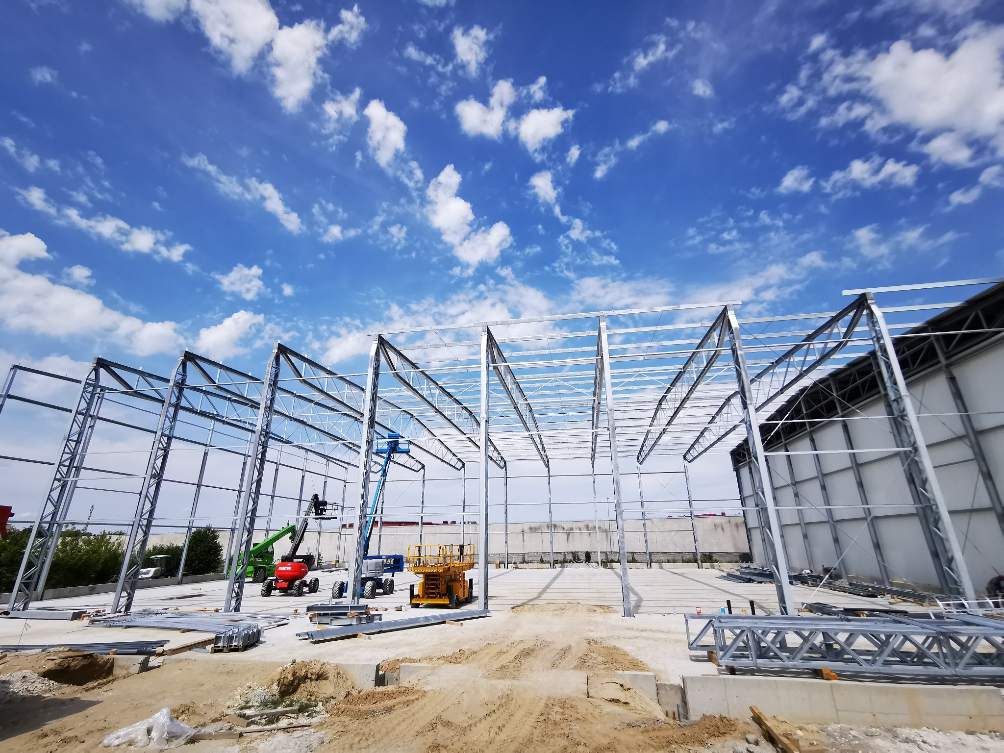 Konstrukcja stalowa hali w trakcie montażu, widok z dołu na niebieskie niebo z chmurami. Na placu budowy obecne są podnośniki i elementy konstrukcyjne.