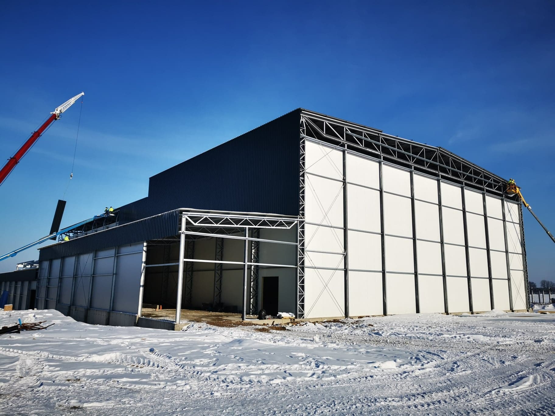 Montaż hali z płyt warstwowych w zimowej scenerii. Widoczny dźwig i pracownicy na wysokości. Częściowo ośnieżony teren budowy, błękitne niebo.