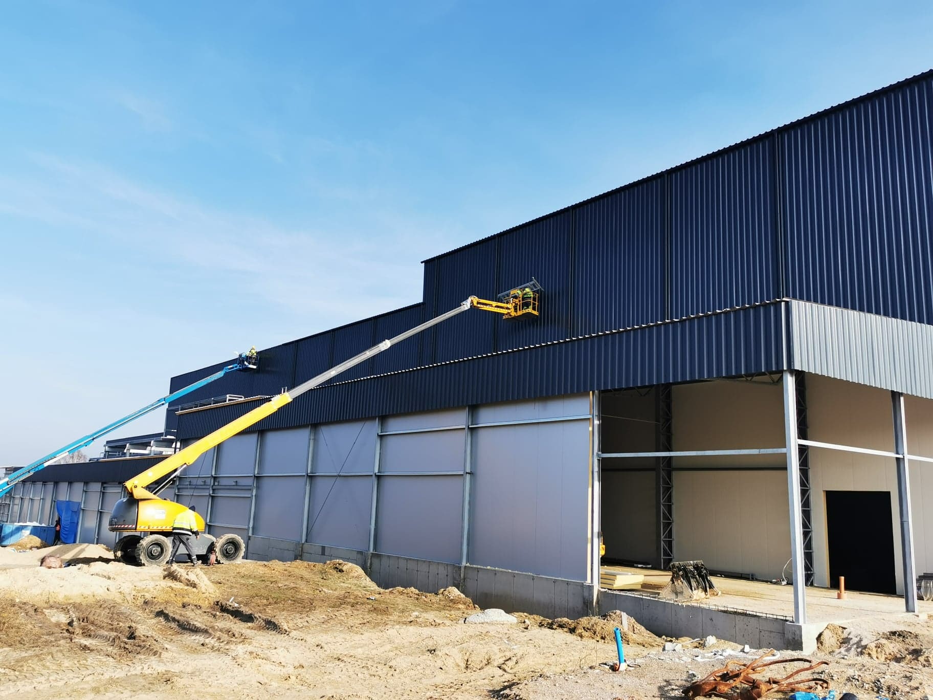 Montaż ciemnej elewacji hali z płyt warstwowych. Dwie platformy robocze z pracownikami na tle błękitnego nieba. Widoczne stalowe elementy konstrukcyjne i teren budowy.