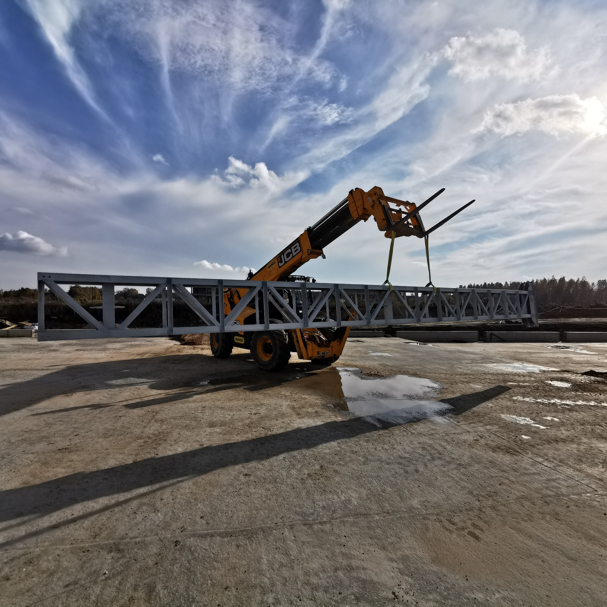 Żółty wózek widłowy JCB podnosi stalową kratownicę na placu budowy. Błękitne niebo z chmurami w tle. Widoczne kałuże i cień wózka.