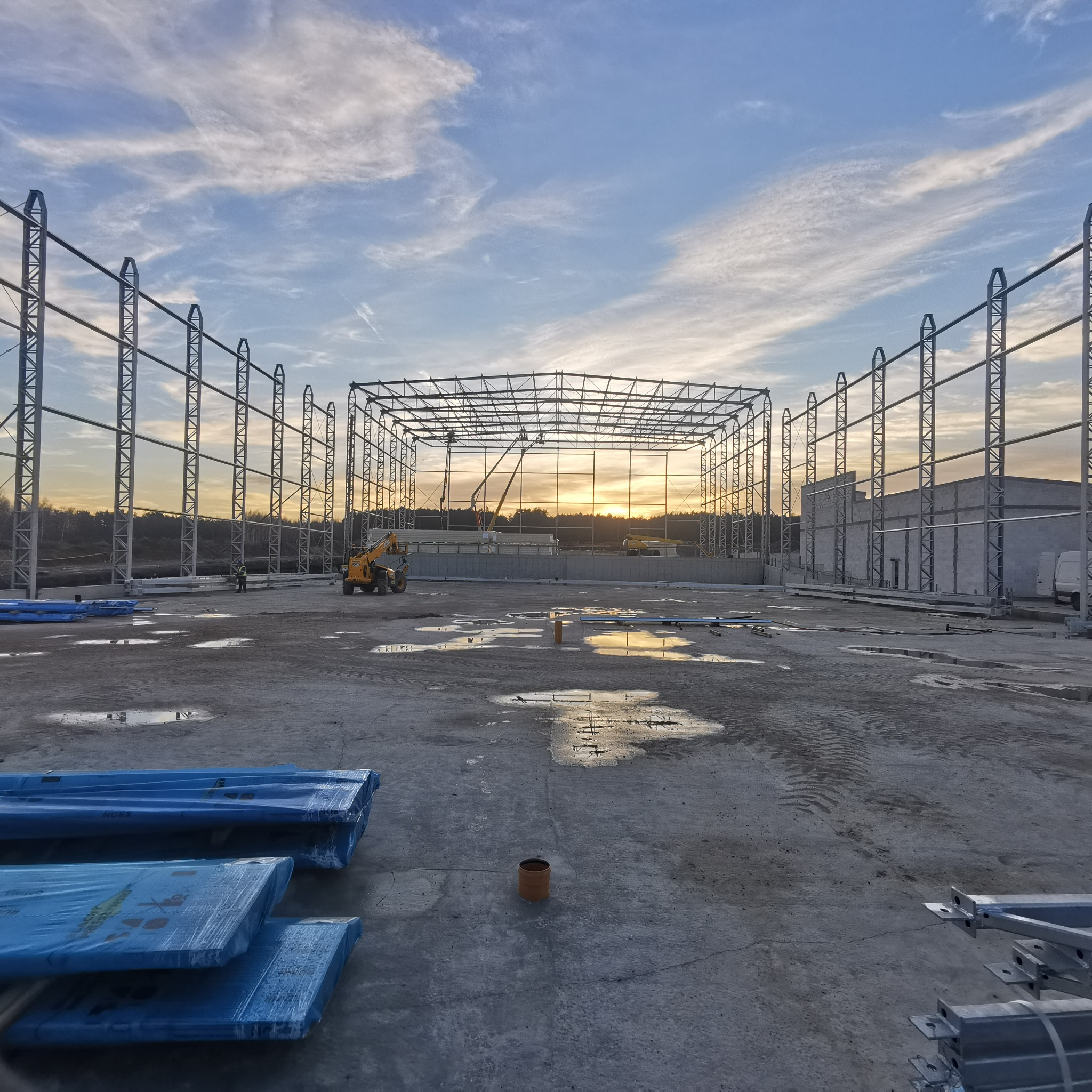 Konstrukcja stalowa hali w trakcie budowy, widoczny szkielet z metalowych belek na tle zachodzącego słońca. Na placu budowy leżą płyty warstwowe w niebieskiej folii.