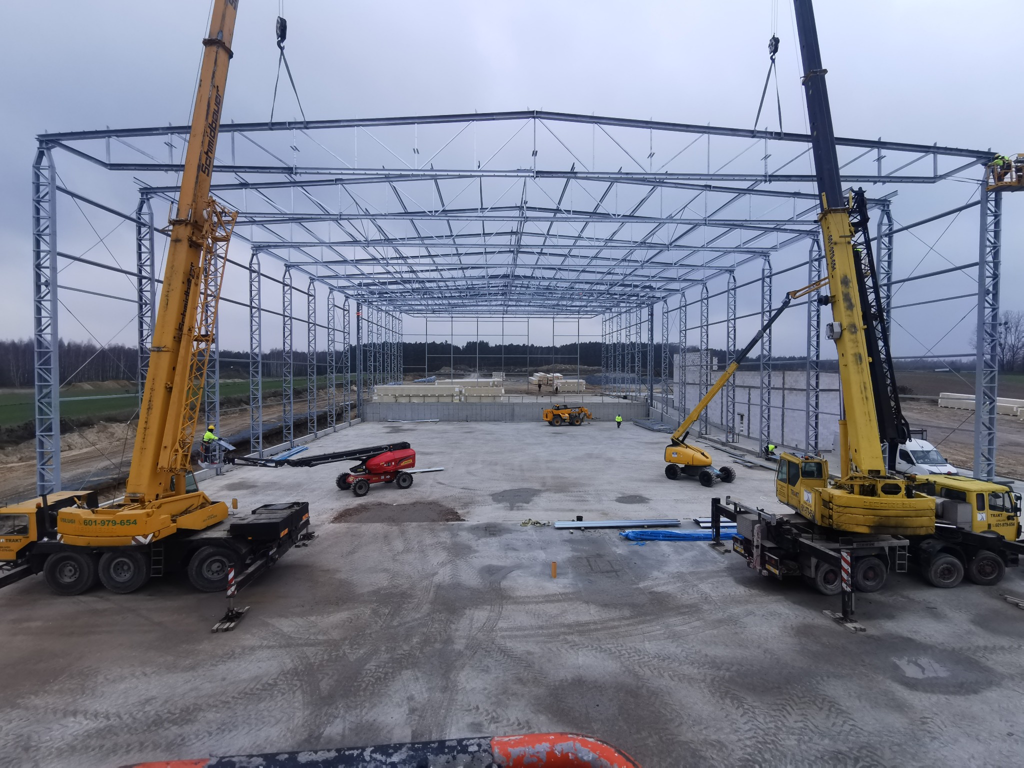 Montaż stalowej konstrukcji hali z użyciem dwóch dźwigów. Widoczny szkielet hali, maszyny budowlane i pracownicy na wysokości. Etap budowy, teren budowy.