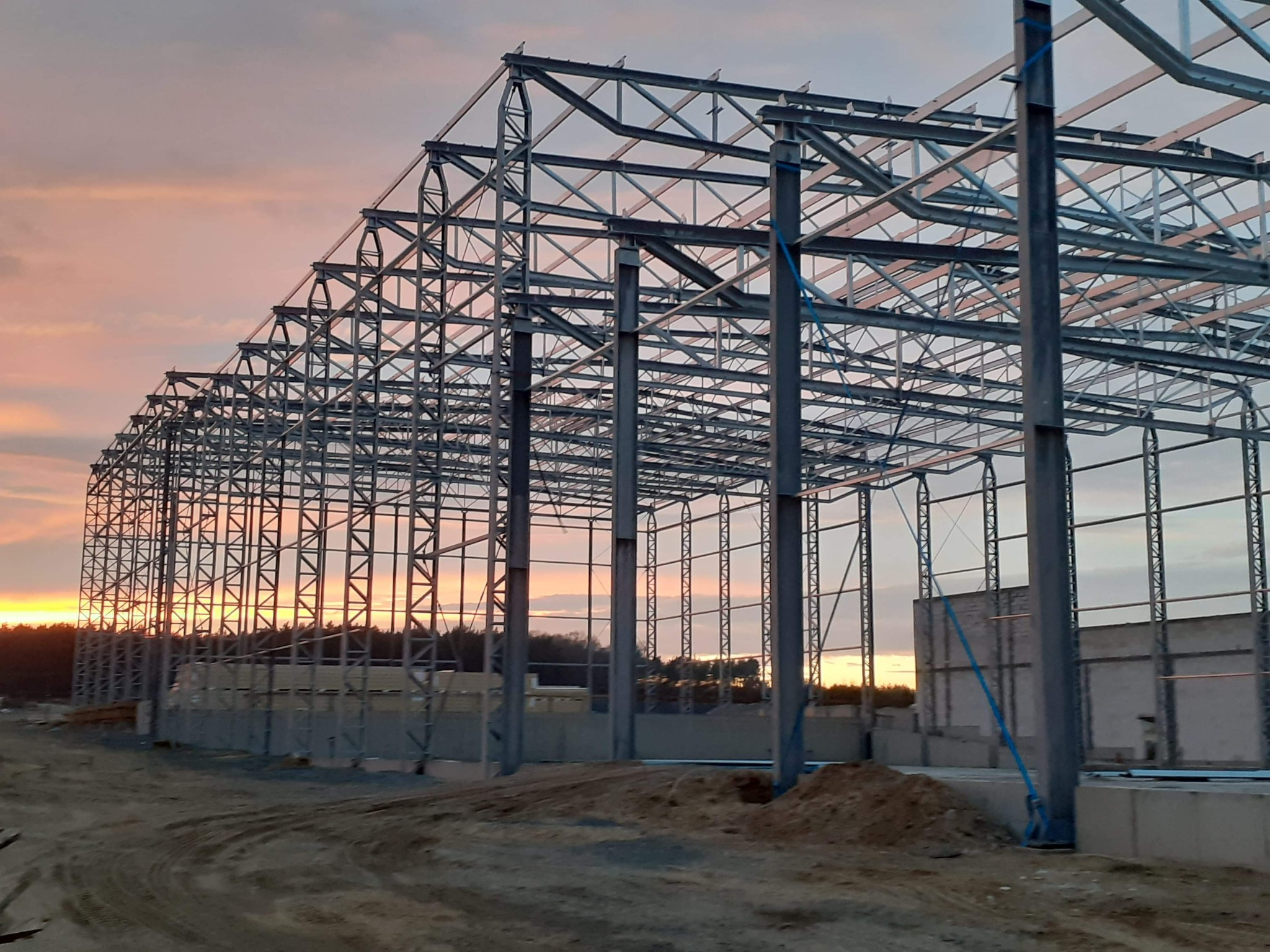 Konstrukcja stalowa hali w trakcie budowy, widoczna na tle zachodzącego słońca. Stalowe elementy tworzą szkielet budynku, z widocznymi połączeniami i wzmocnieniami.