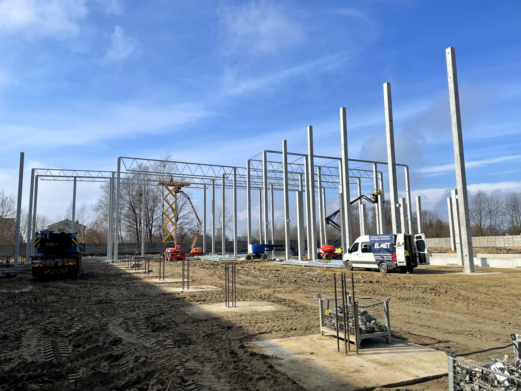 Budowa hali stalowej w trakcie montażu konstrukcji. Widoczne słupy, dźwigi oraz ekipa montażowa. Na pierwszym planie teren budowy, w tle błękitne niebo z chmurami.