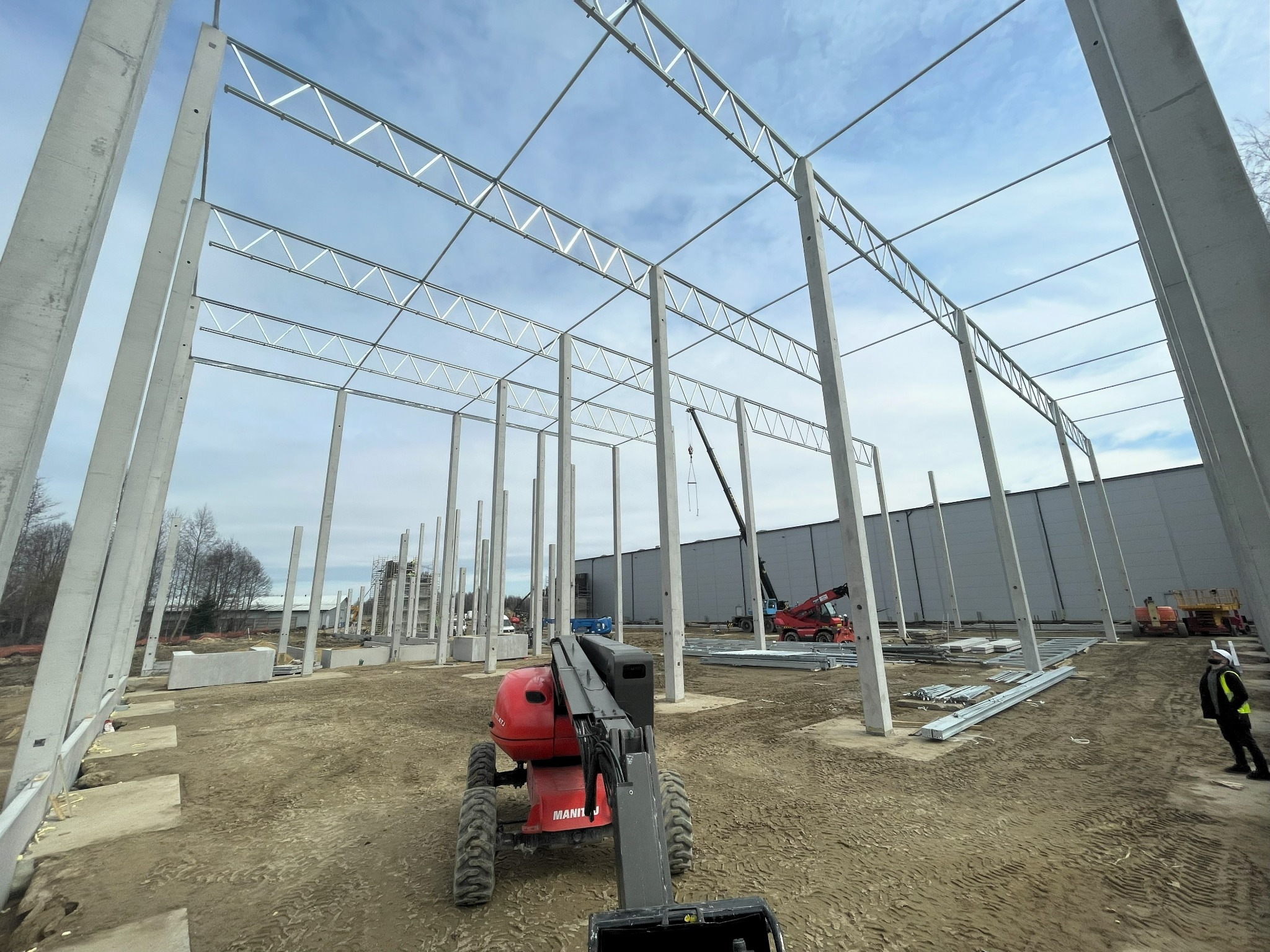 Wznoszenie szkieletu hali stalowej z użyciem podnośnika. Widoczne betonowe słupy i metalowe elementy konstrukcyjne. Częściowo ukończona budowa, teren budowy.