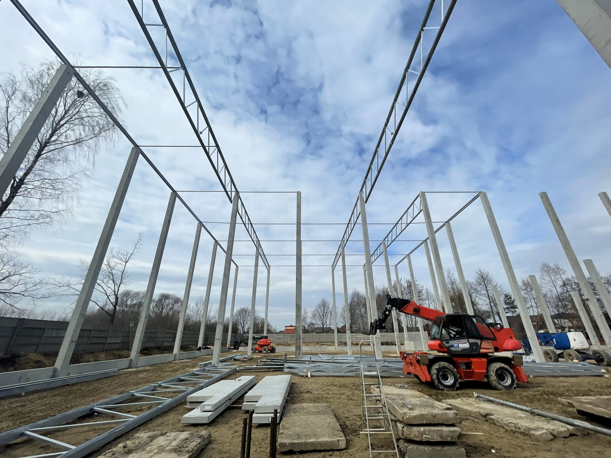 Wnętrze budowanej hali z betonu i stali, widok z poziomu gruntu na konstrukcję, z czerwonym podnośnikiem teleskopowym w tle i elementami szkieletu na ziemi.