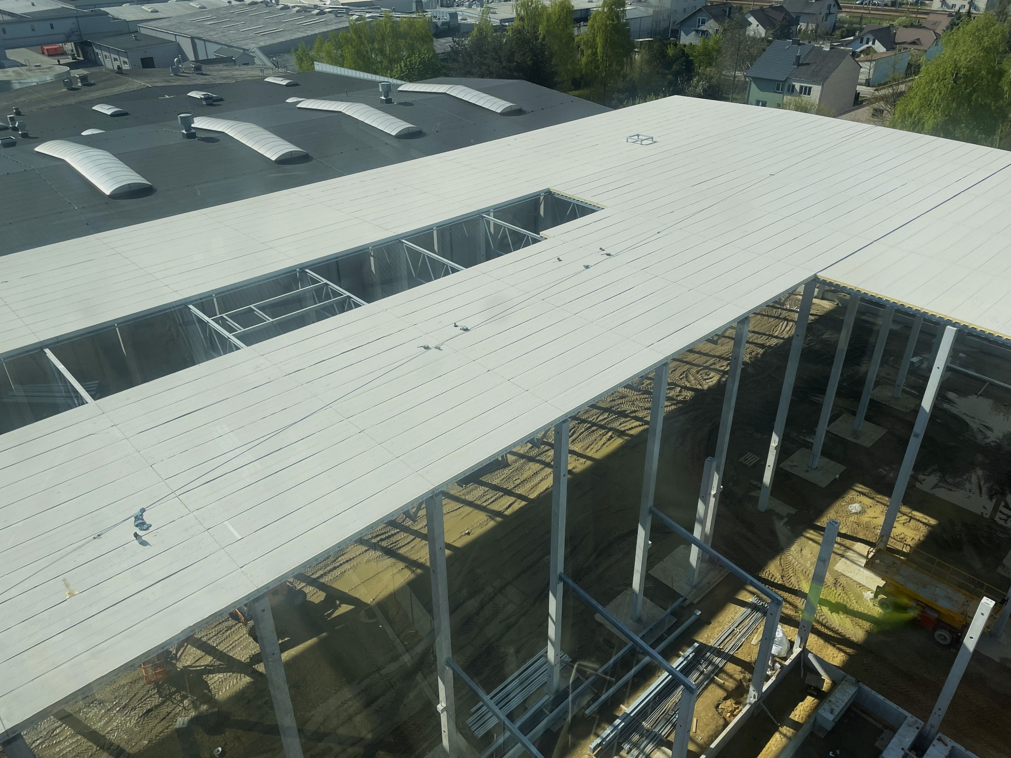 Wysoki kąt widoku na budowę hali z widoczną konstrukcją stalową, częściowo pokrytą białymi panelami. W tle budynki mieszkalne i przemysłowe. Widoczne otwory w dachu.