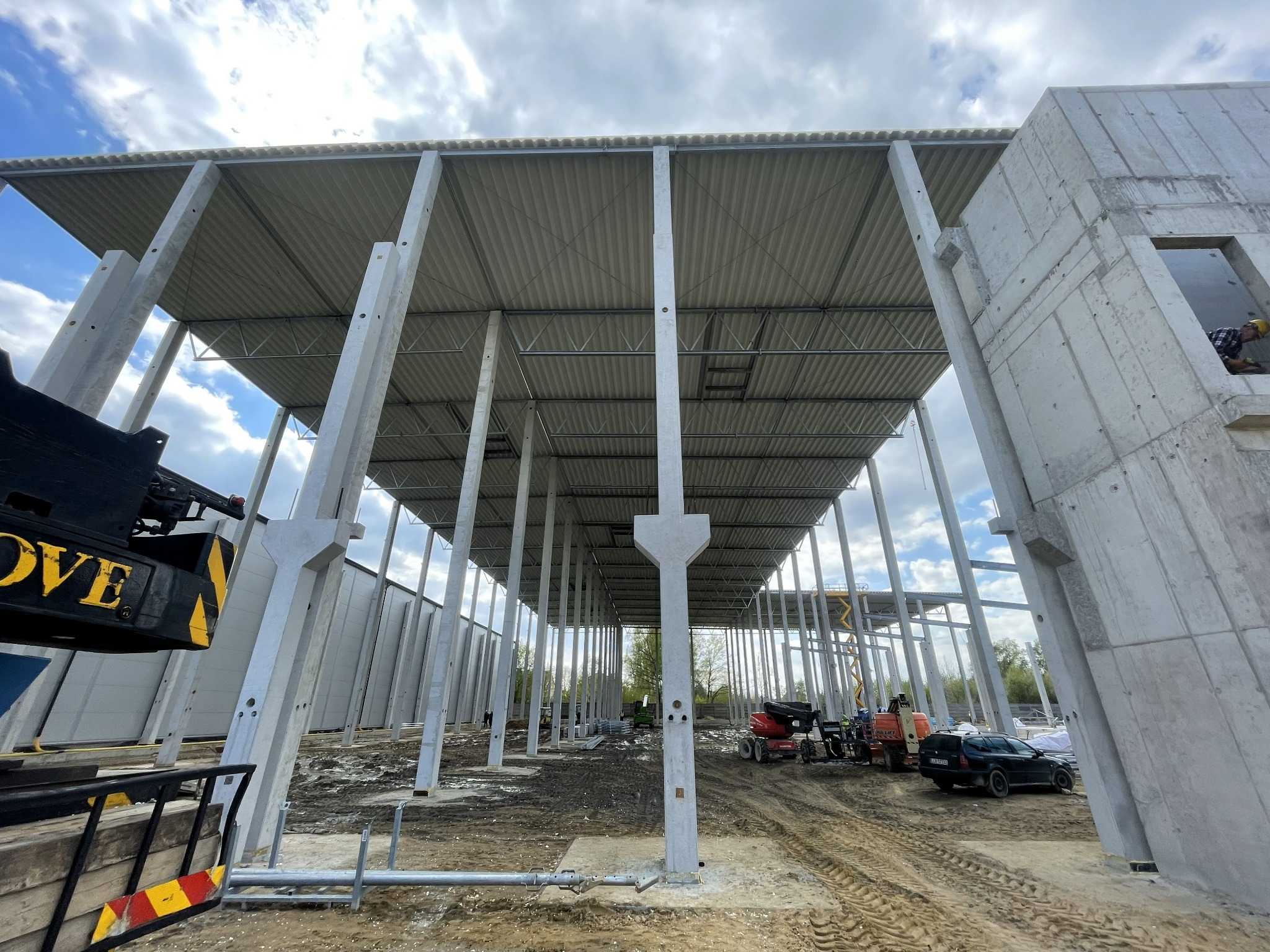 Wnętrze hali w budowie: betonowe słupy wspierają blaszany dach. Na placu budowy sprzęt i samochód osobowy. Widok z dołu na konstrukcję pod chmurami.