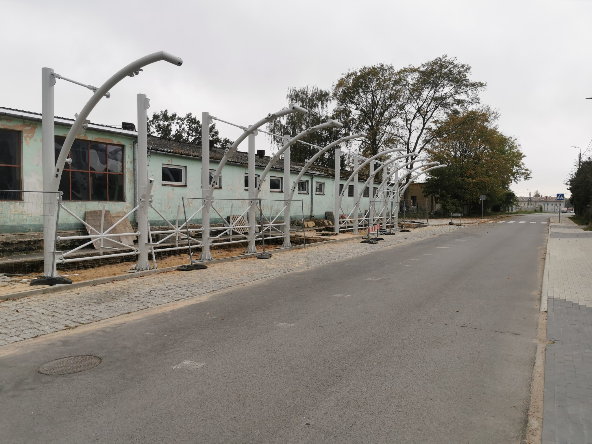Montaż metalowej konstrukcji nad chodnikiem przy budynku. Widoczne słupy i łukowe elementy. Prace w toku, teren zabezpieczony siatką. Dzień pochmurny.