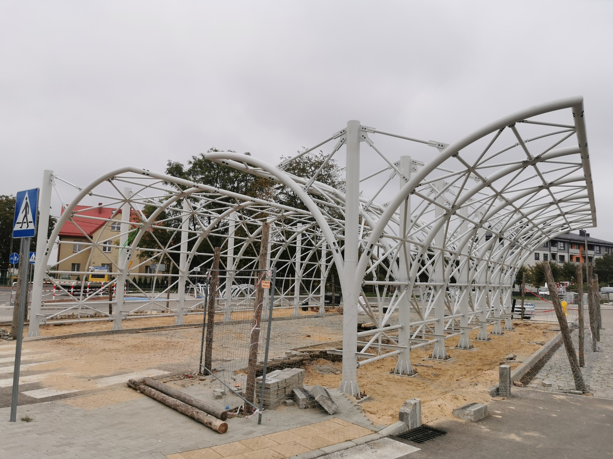 Biała, łukowa konstrukcja stalowa w trakcie budowy, widoczna na tle miejskiego krajobrazu, częściowo ogrodzona siatką, z elementami infrastruktury drogowej.