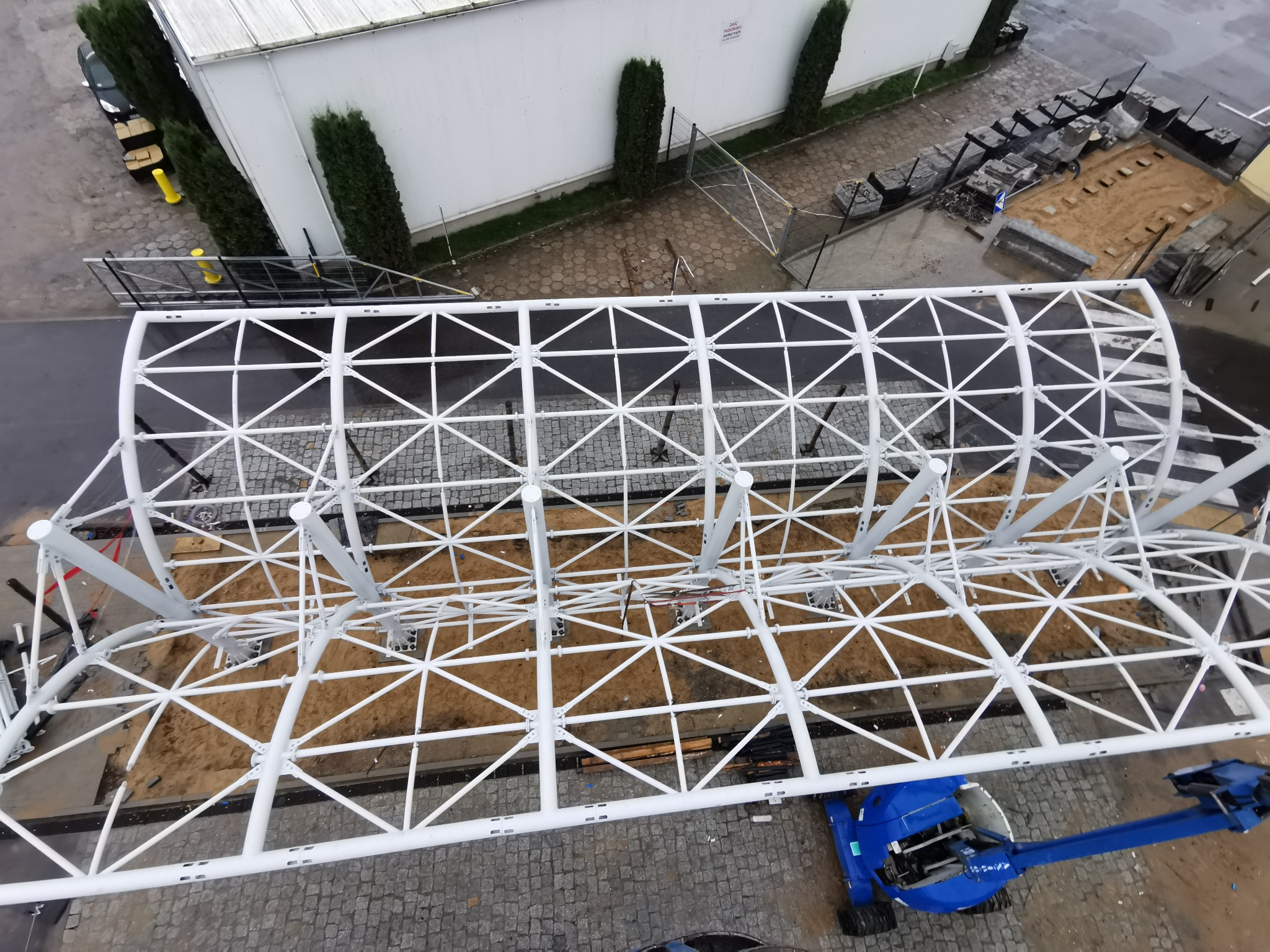 Widok z góry na białą konstrukcję stalową w Wiśniewie, prawdopodobnie zadaszenie, z użyciem podnośnika. Konstrukcja na placu z kostki brukowej.