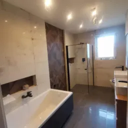 Nowoczesna łazienka z wanną w zabudowie i kabiną prysznicową typu walk-in, wykończona płytkami w odcieniach beżu i brązu z elementami dekoracyjnymi na ścianie.