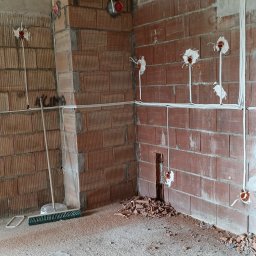 Michał Gumulski - Instalacja elektryczna w surowym wnętrzu: kable biegnące po ceglanych ścianach i pod sufitem, puszki elektryczne w ścianach, gruz na podłodze, miotła w rogu.