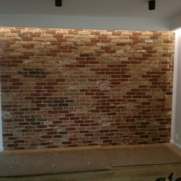 Michał Gumulski - Ściana z cegły dekoracyjnej w neutralnych barwach, oświetlona od góry, z widocznymi elementami podłogi i sufitu. Wnętrze w trakcie wykańczania.