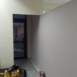 Michał Gumulski - Świeżo pomalowane ściany w odcieniach beżu i szarości, z widocznym wejściem i workiem na śmieci. W tle widoczny fragment ulicy przez szklane drzwi.