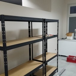 Michał Gumulski - Metalowy regał magazynowy z drewnianymi półkami w jasnym pomieszczeniu. Widoczne narzędzia na jednej z półek i biała lodówka w tle.