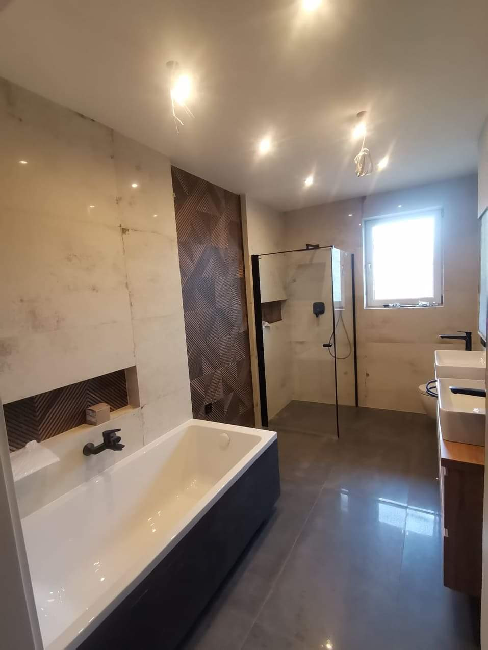 Nowoczesna łazienka z wanną w zabudowie i kabiną prysznicową typu walk-in, wykończona płytkami w odcieniach beżu i brązu z elementami dekoracyjnymi na ścianie.