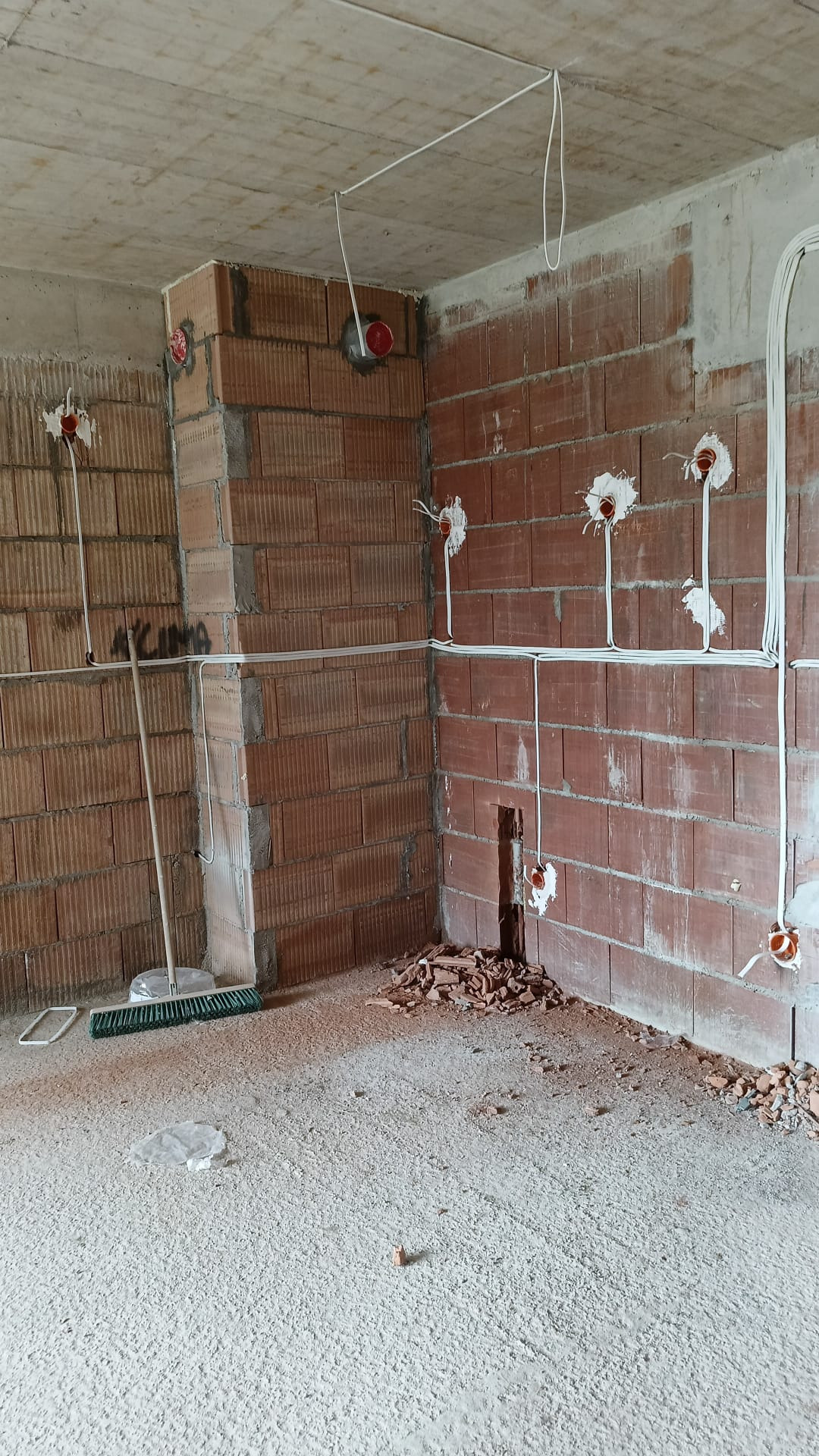 Instalacja elektryczna w surowym wnętrzu: kable biegnące po ceglanych ścianach i pod sufitem, puszki elektryczne w ścianach, gruz na podłodze, miotła w rogu.