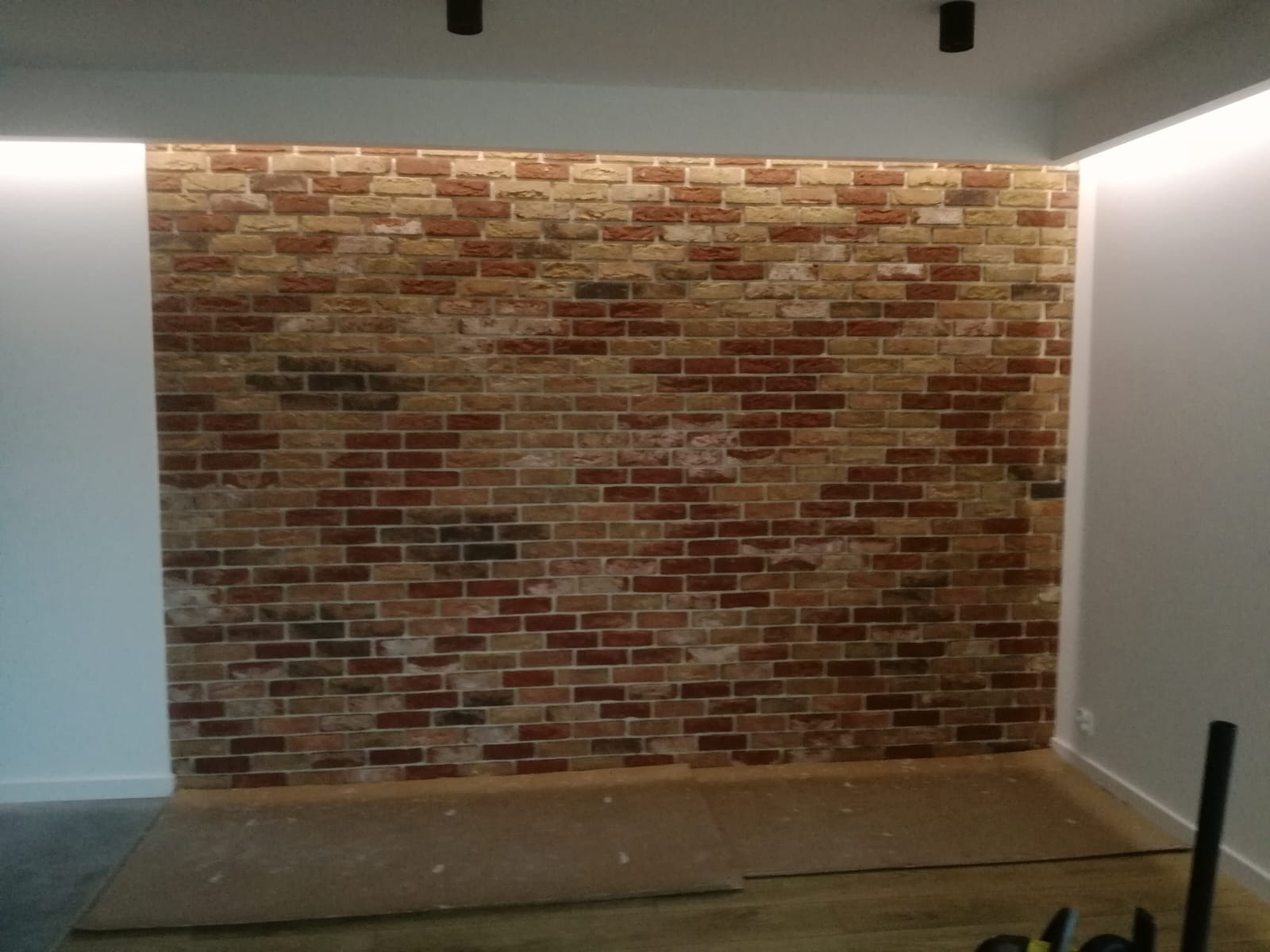 Ściana z cegły dekoracyjnej w neutralnych barwach, oświetlona od góry, z widocznymi elementami podłogi i sufitu. Wnętrze w trakcie wykańczania.