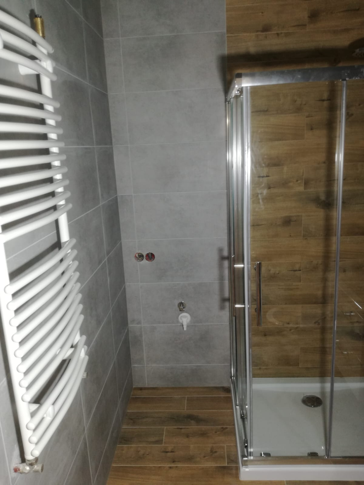 Szara łazienka z grzejnikiem drabinkowym, kabiną prysznicową z drewnopodobnymi płytkami. Widoczne przyłącza wodne i odpływ. Ukończony remont.