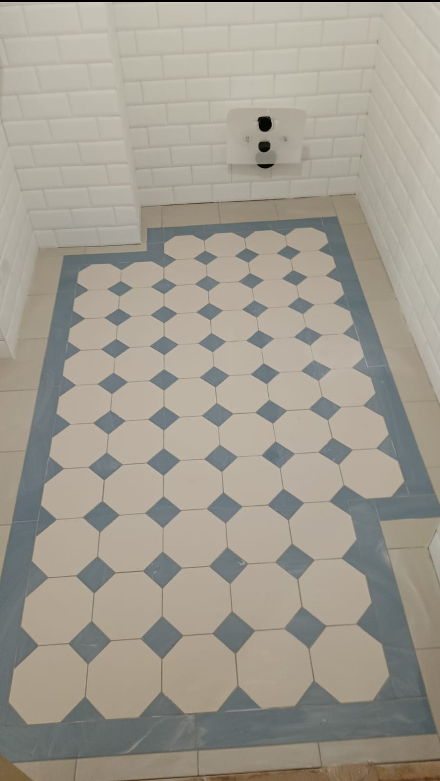 Ukończona podłoga w łazience z białych i niebieskich płytek o geometrycznym wzorze, z białymi płytkami na ścianach i podwieszanym WC.