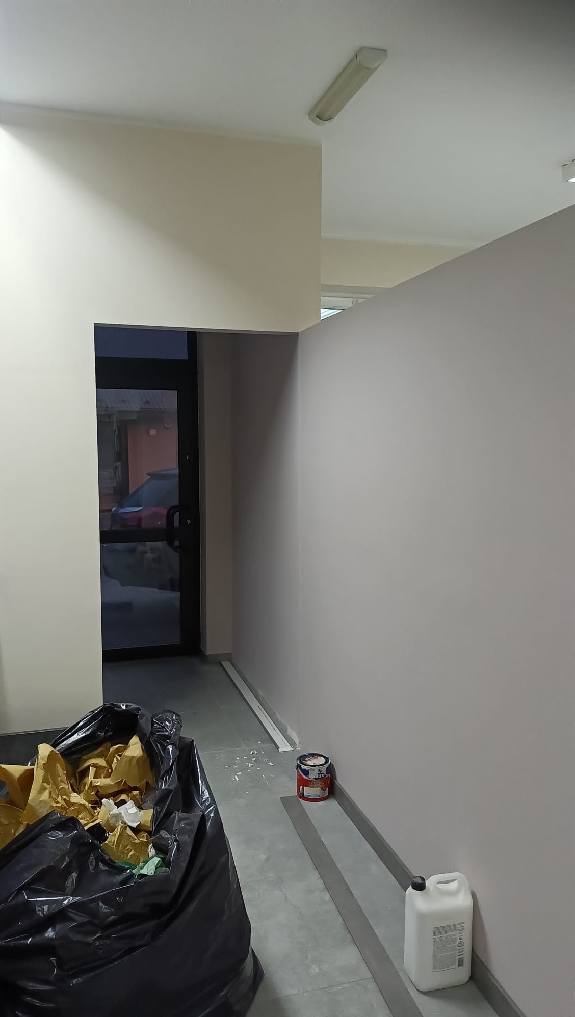 Świeżo pomalowane ściany w odcieniach beżu i szarości, z widocznym wejściem i workiem na śmieci. W tle widoczny fragment ulicy przez szklane drzwi.