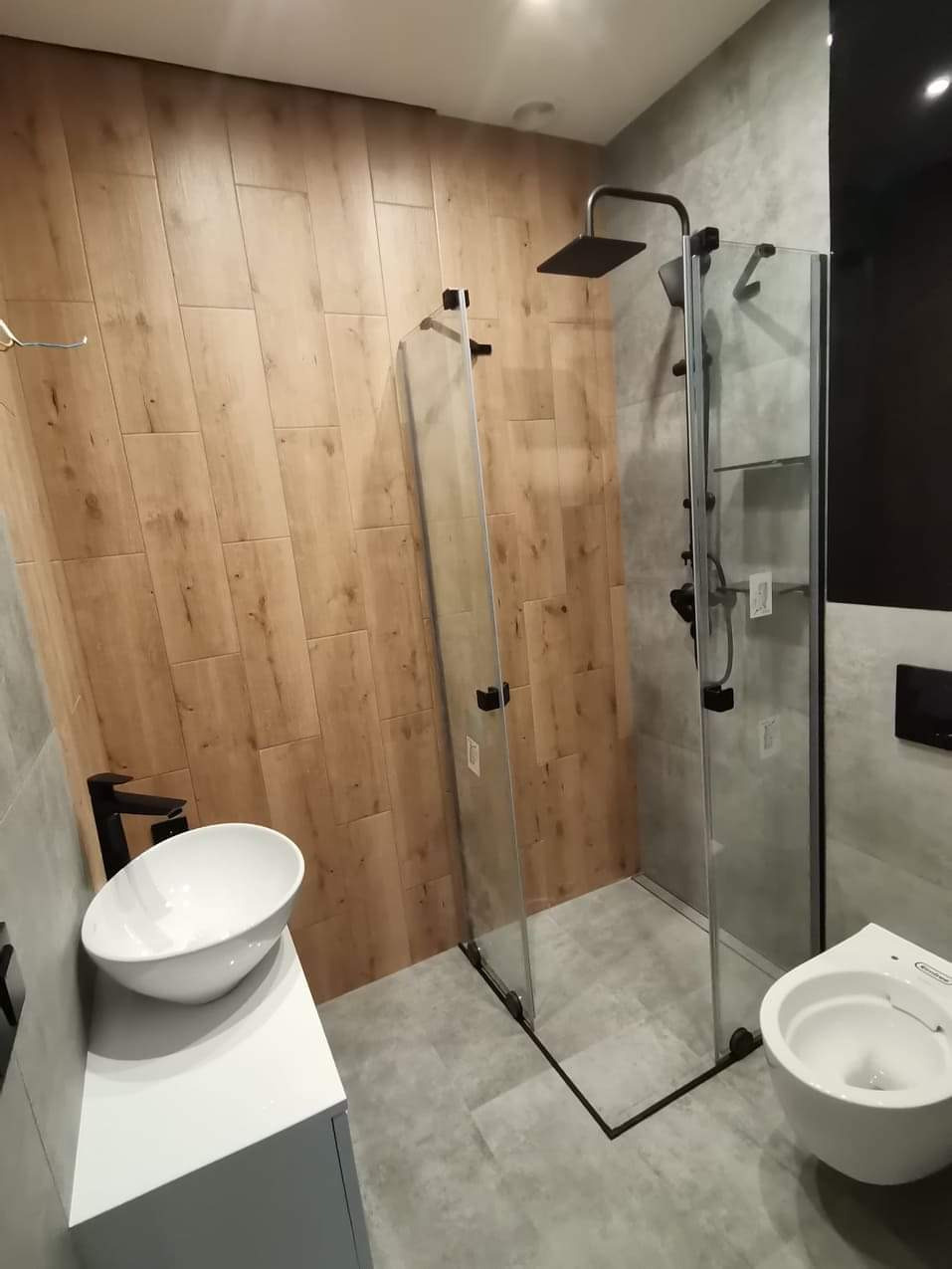 Nowoczesna łazienka z kabiną typu walk-in, drewnopodobnymi płytkami na ścianie i minimalistyczną armaturą w czarnym macie. Umywalka nablatowa i szara podłoga.
