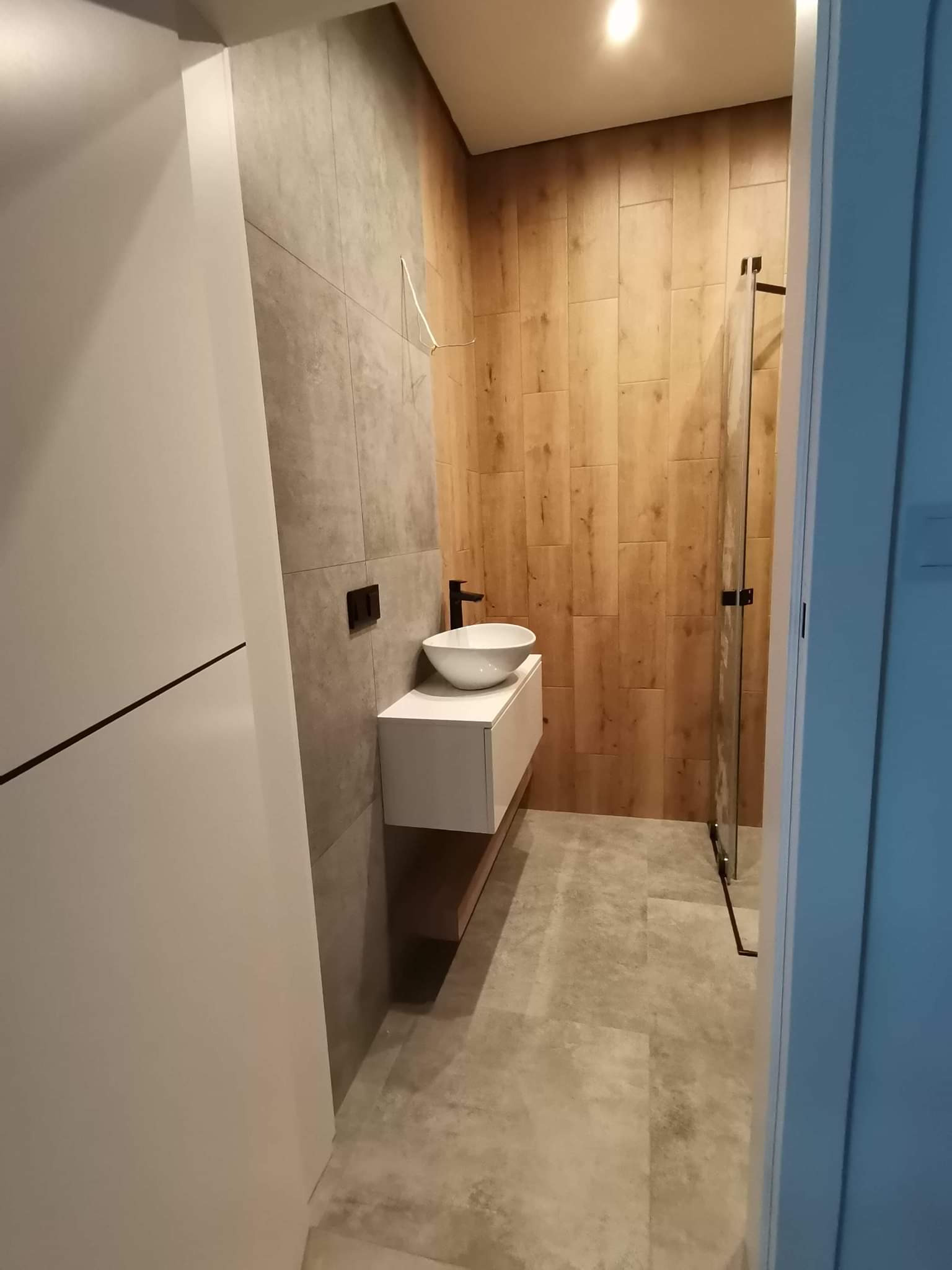 Nowoczesna łazienka z drewnopodobnymi i szarymi płytkami, białą umywalką nablatową i kabiną prysznicową z czarnymi detalami. Minimalistyczny design i ciepłe oświetlenie.