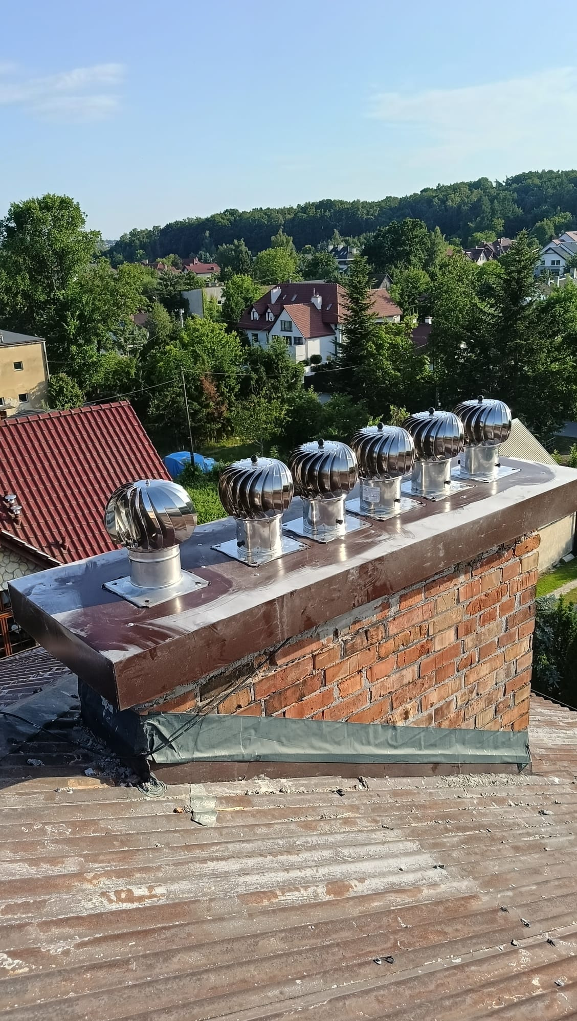 Ceglany komin z zamontowanymi, obrotowymi nasadami wentylacyjnymi. W tle zabudowa jednorodzinna i zieleń.