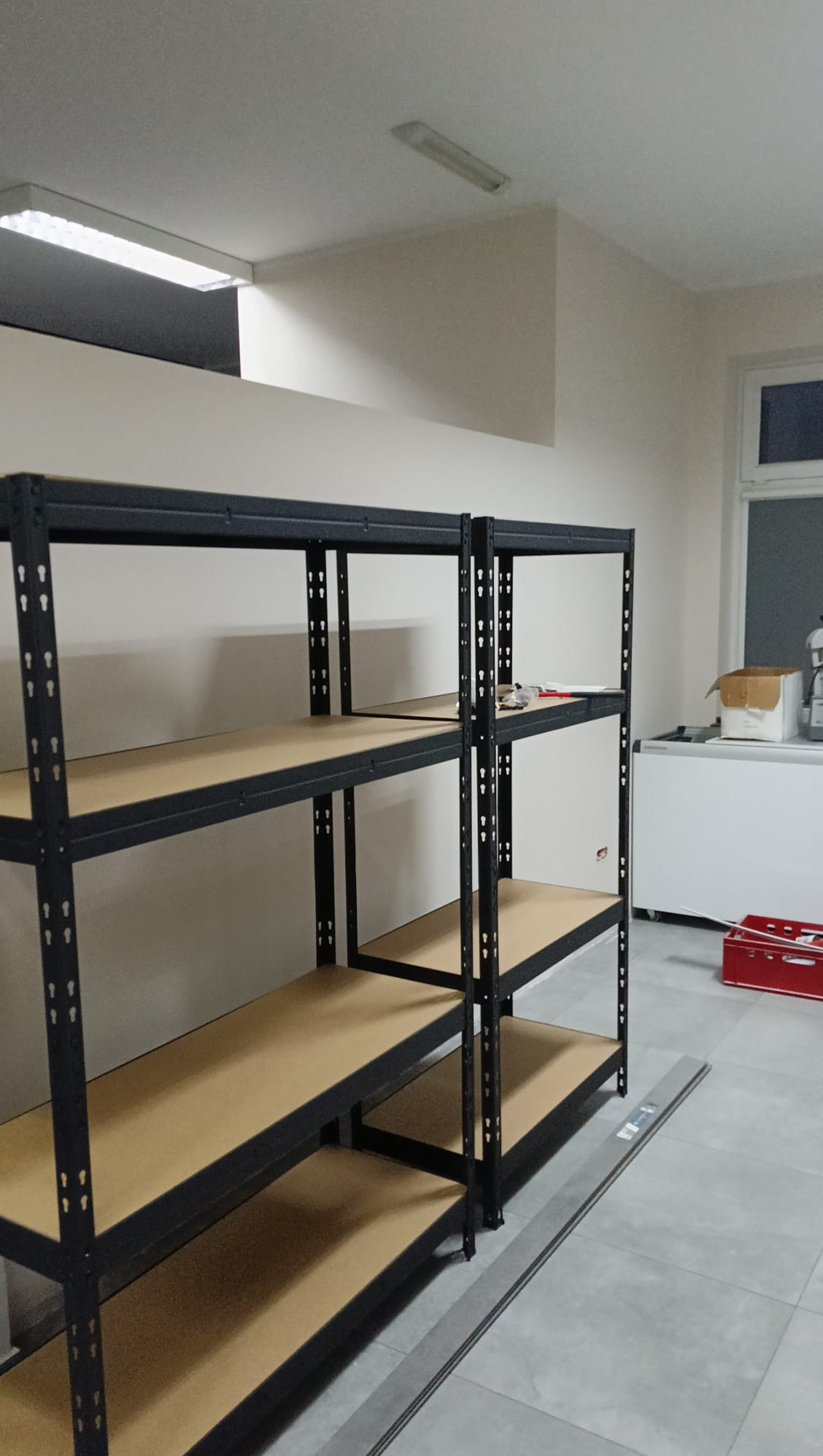 Metalowy regał magazynowy z drewnianymi półkami w jasnym pomieszczeniu. Widoczne narzędzia na jednej z półek i biała lodówka w tle.