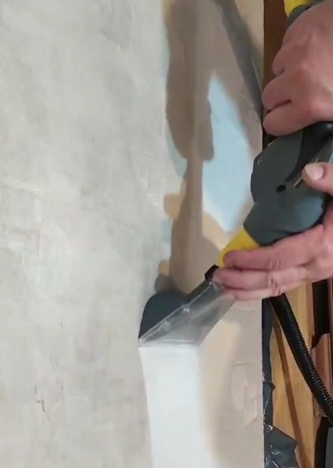 Czyszczenie tapicerki beżowego krzesła za pomocą parownicy z przezroczystą końcówką i żółtą dyszą. Widoczne dłonie operatora i fragment podłogi.