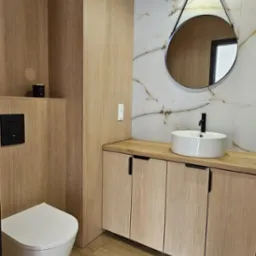 Nowoczesna łazienka z drewnopodobnymi płytkami, wiszącą miską WC, szafką pod umywalką z blatem i okrągłym lustrem z oświetleniem.