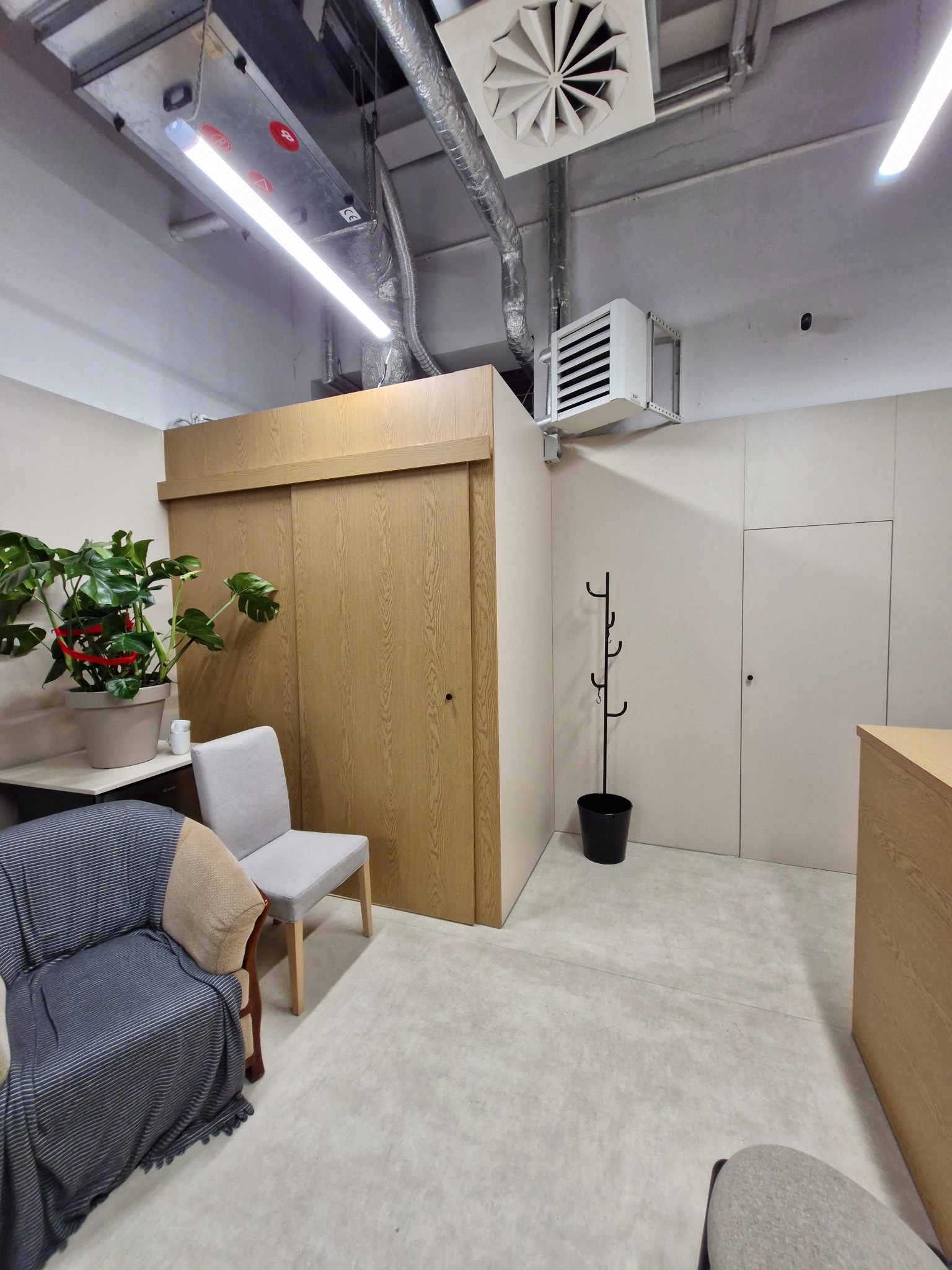 Jasne wnętrze z drewnianą szafą przesuwną, rośliną doniczkową i krzesłami. Widoczne instalacje wentylacyjne pod sufitem. Minimalistyczny design.