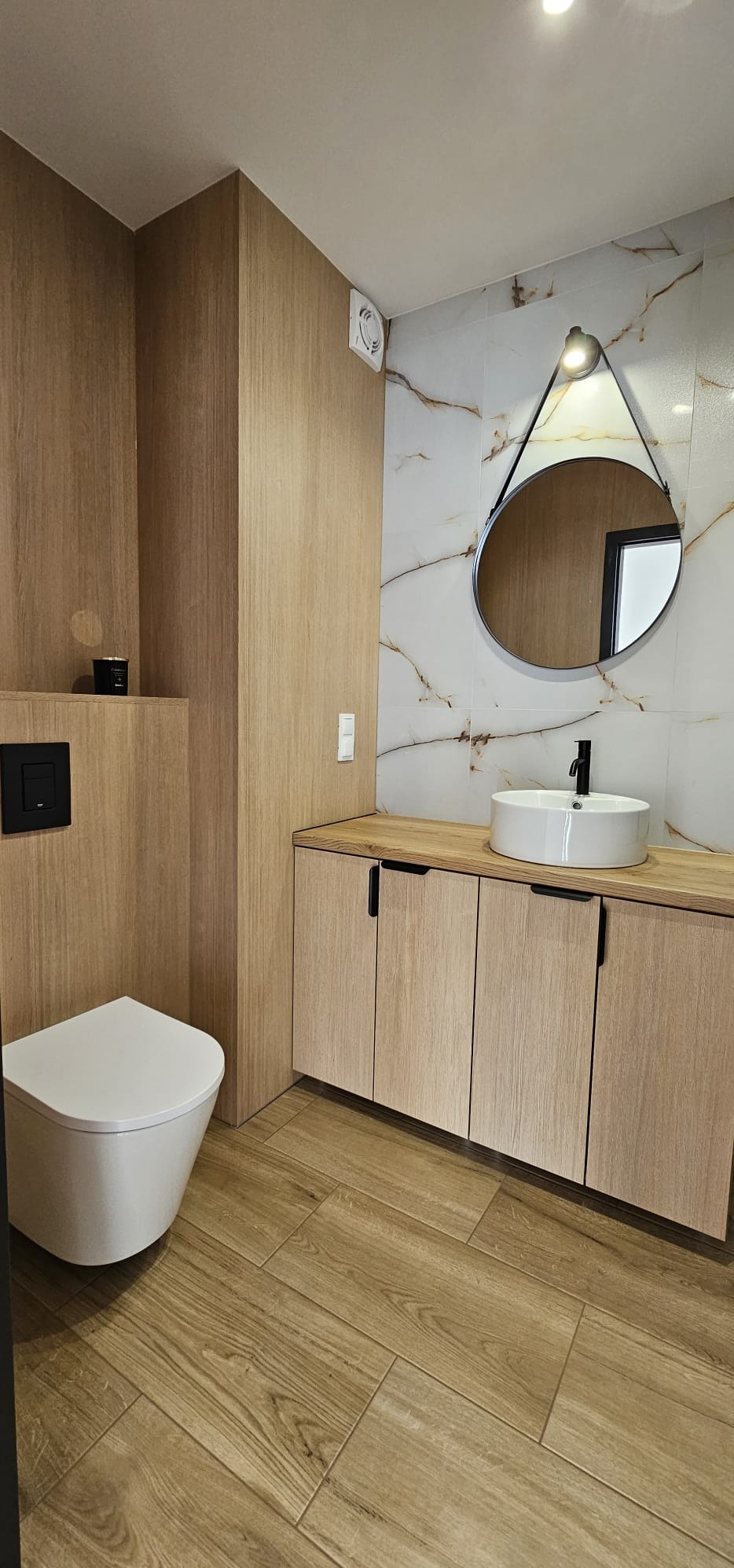 Nowoczesna łazienka z drewnopodobnymi płytkami, wiszącą miską WC, szafką pod umywalką z blatem i okrągłym lustrem z oświetleniem.