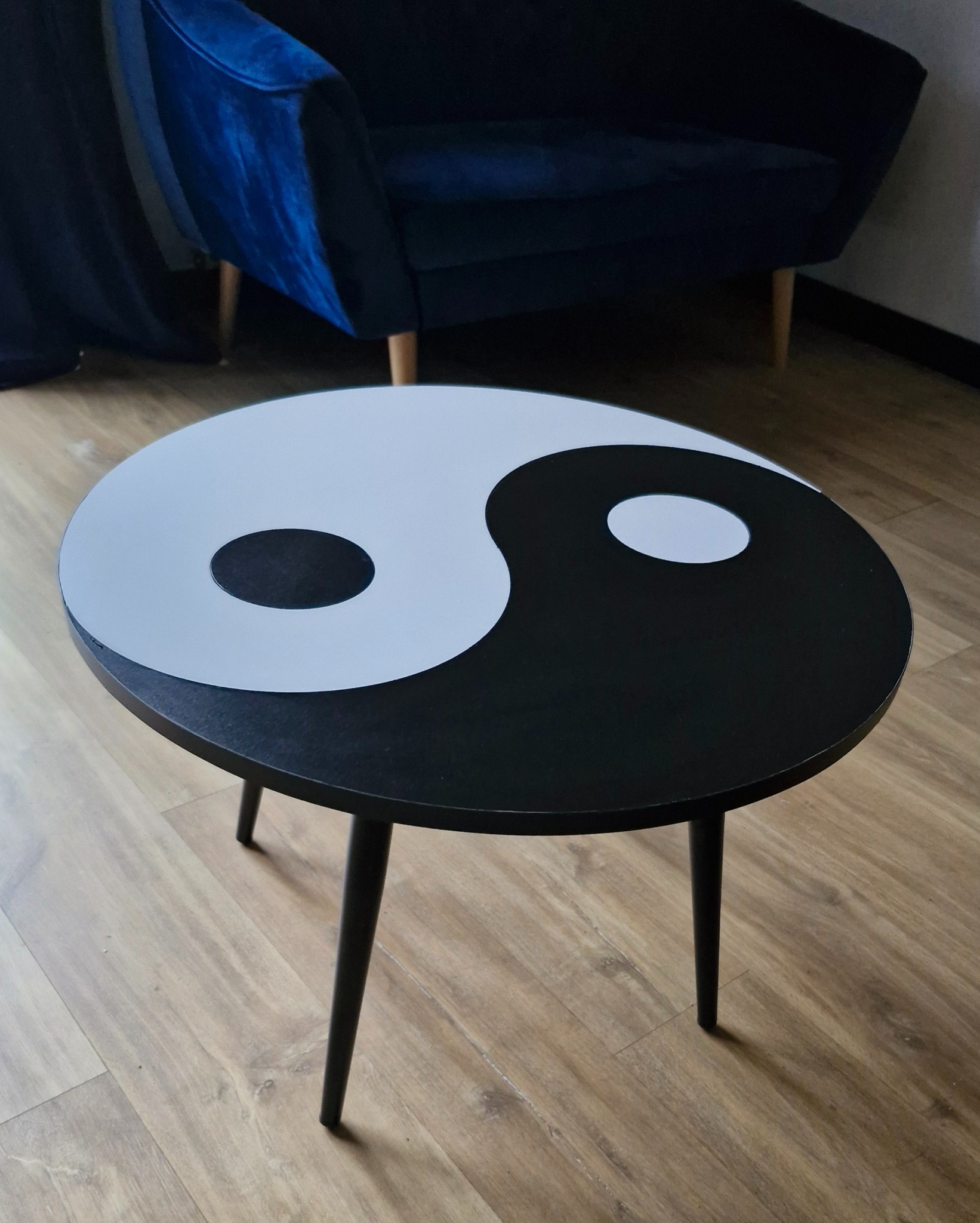 Okrągły stolik kawowy z blatem w stylu Yin Yang, ustawiony na drewnianej podłodze, z fragmentem granatowego fotela w tle. Minimalistyczny design.