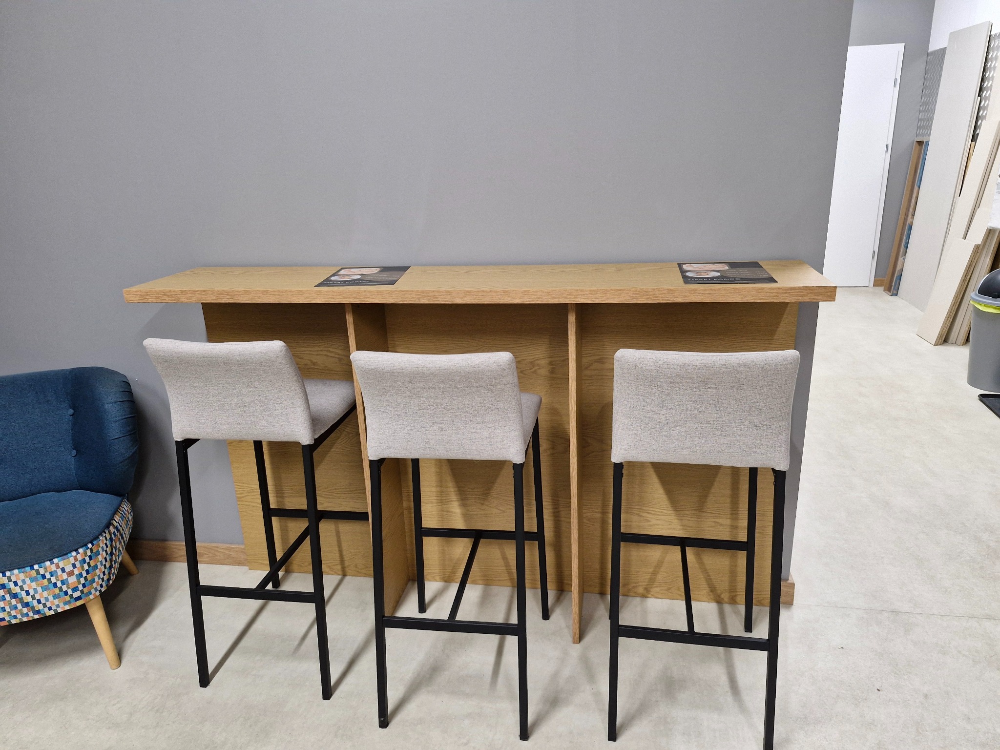 Wysoki, drewniany stół barowy z trzema krzesłami w nowoczesnym wnętrzu. Blat z jasnego drewna, czarne nogi krzeseł. W tle szara ściana i elementy wyposażenia.