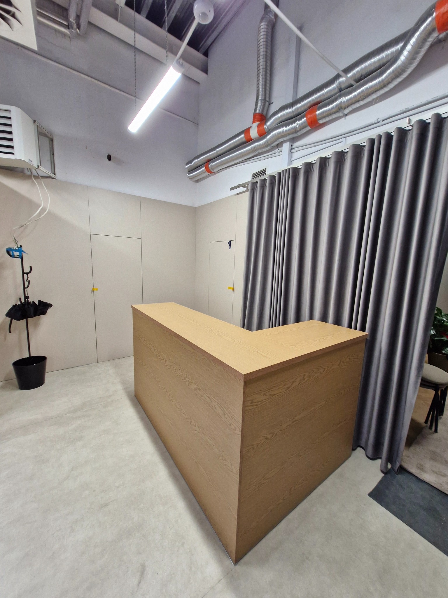 Lada recepcyjna z drewnianym wykończeniem w jasnym wnętrzu, z widocznymi elementami instalacji sufitowej i zasłonami w tle. Minimalistyczny design.