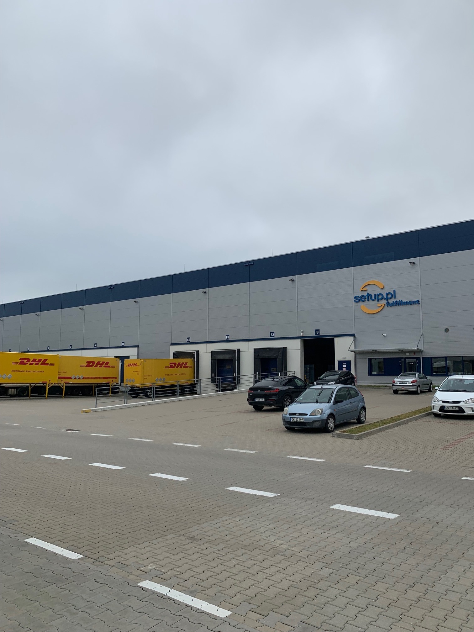 Magazyn firmy Setup.pl Fulfillment z żółtymi ciężarówkami DHL przy rampach. Szaro-błękitny budynek magazynowy z numerami bram. Parking przed budynkiem.